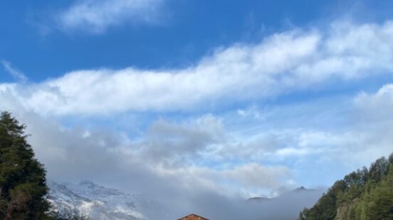 A wooden cabin with a snow-capped mountain backdrop under a blue sky with white clouds.