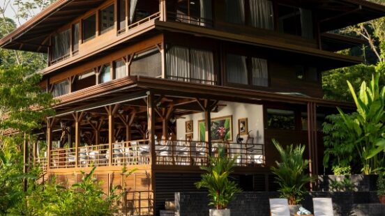 A luxurious wooden house with balconies by a pool, surrounded by lush greenery.