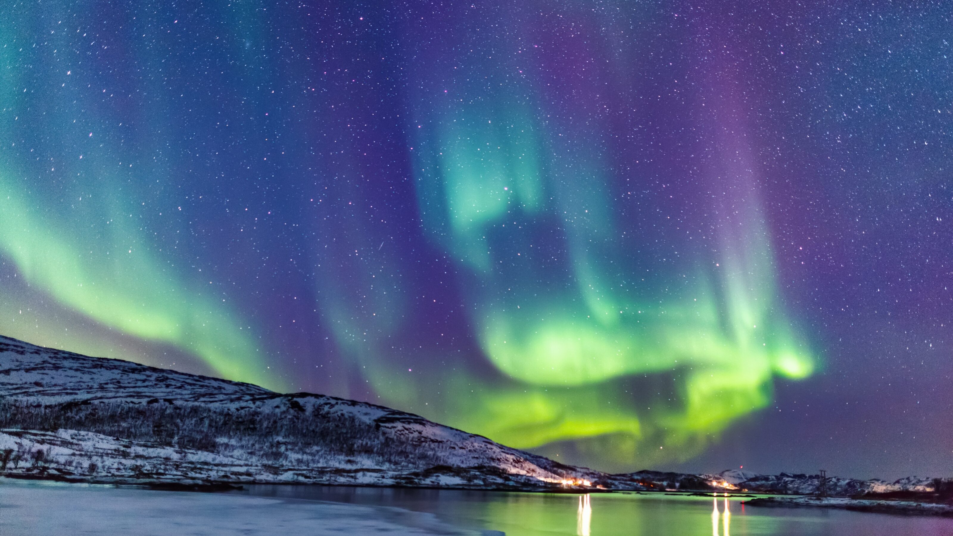 A vivid aurora borealis above a snowy landscape and icy water with stars twinkling in the sky.