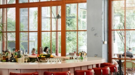 Elegant bar interior with chandelier, red stools, and large windows overlooking greenery.