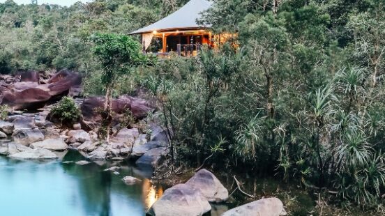 A house with lit interior by a serene river surrounded by forest and rocks at twilight.