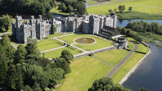 Aerial view of a historic castle with lush gardens beside a river, adjacent to a golf course.
