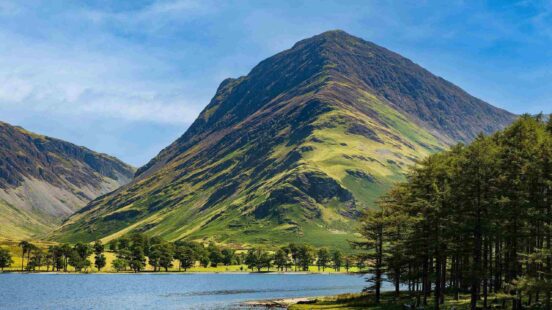 A towering green mountain behind a lake with trees in the foreground under a blue sky.