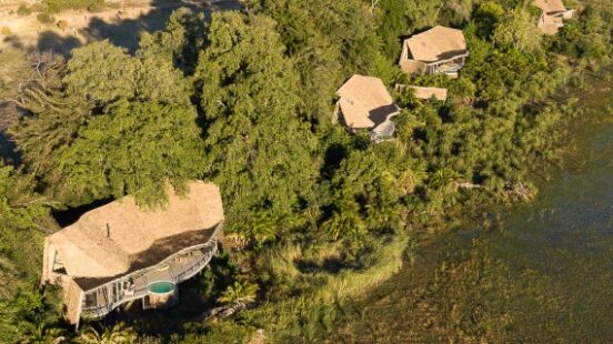 Aerial view of thatched-roof buildings nestled among greenery next to water, with open fields beyond.