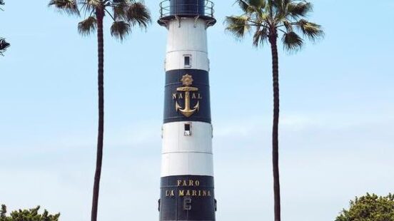 A lighthouse flanked by palm trees under a clear blue sky, with a tiled pathway leading to it.