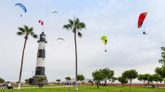 A lighthouse with a Peruvian flag, surrounded by palm trees and paragliders in the sky.