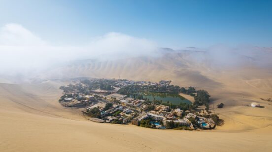 Oasis town with buildings and greenery surrounded by vast sand dunes under a hazy sky.
