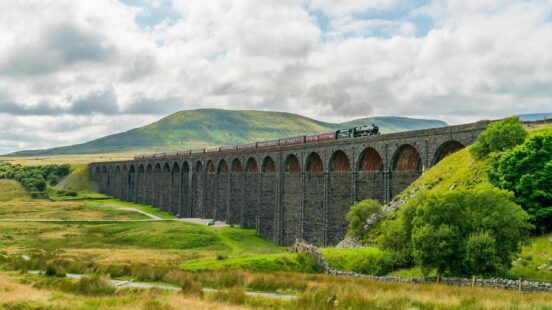 Steam train crossing a large stone viaduct amidst green hills.