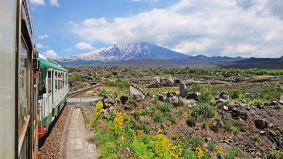 Train traveling through volcanic landscape with snow-capped mountain in the background.
