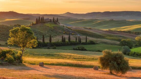 Rolling Tuscan hills at sunset, with hay bales, trees, and a distant villa.