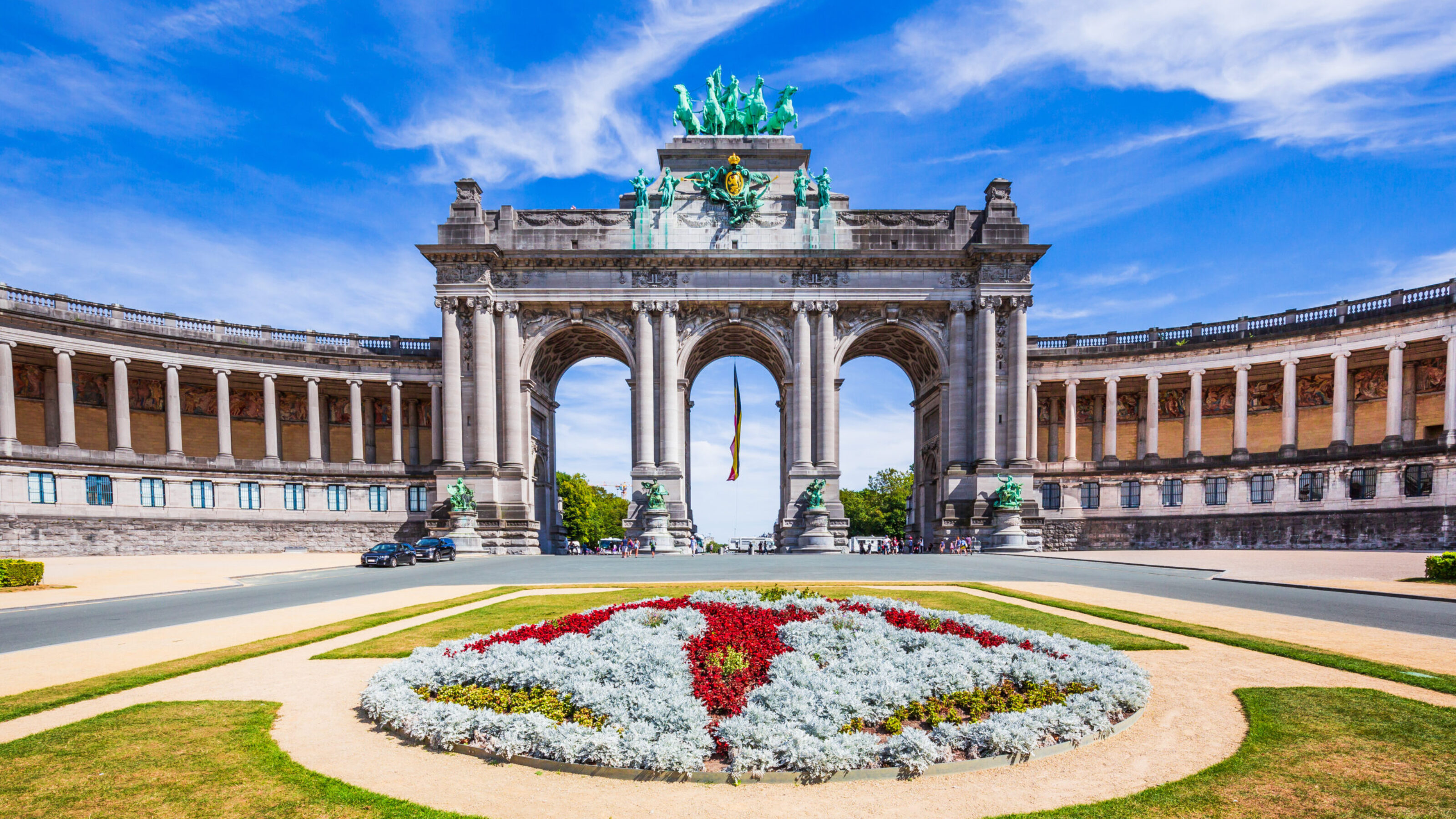 A grand triumphal arch with sculptural details and a colourful floral foreground under a blue sky.