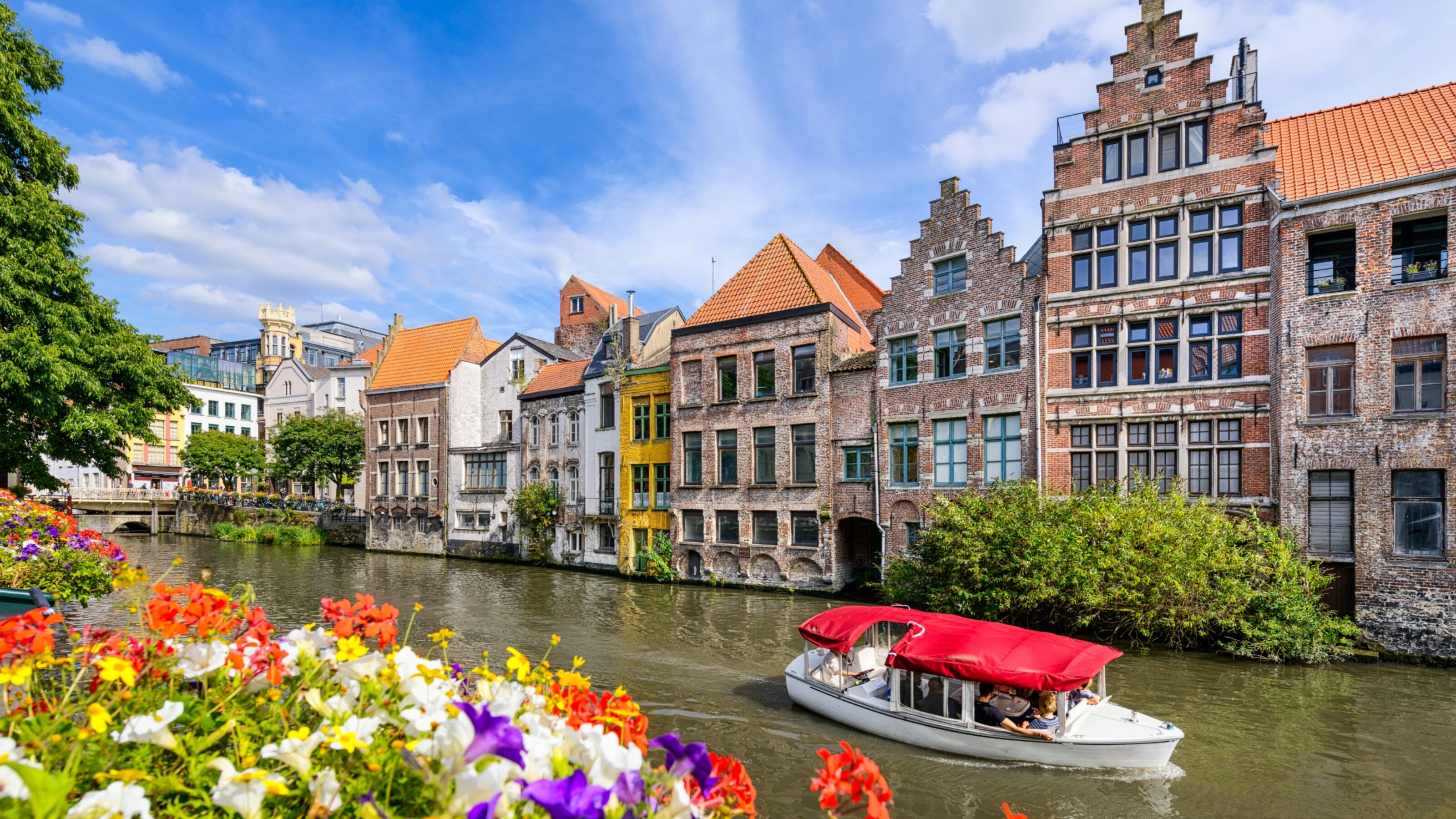 A boat on a canal with historic buildings and colourful flowers in the foreground.