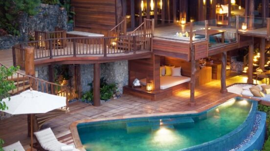 Luxury wooden resort with pool, lit up at twilight nestled in lush greenery.