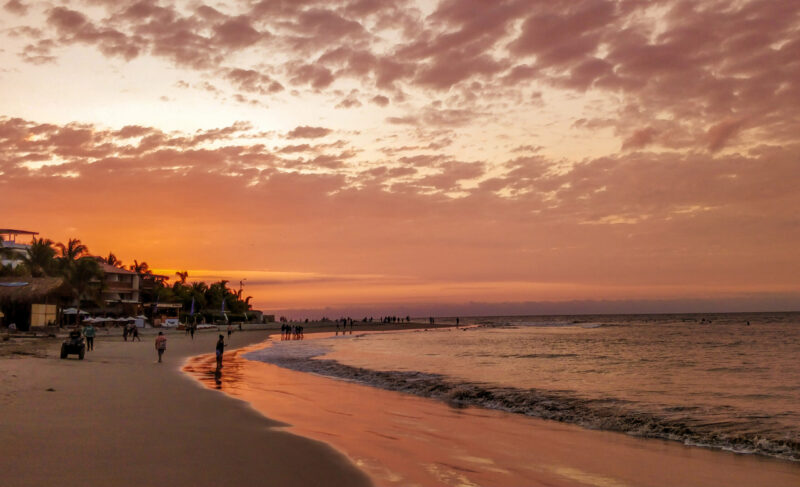 Sunset at the beach with people, reflecting water, and clouds in the sky.