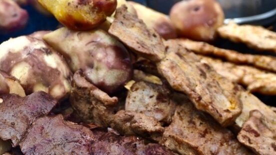 Stack of grilled meats on skewers with pots of food in the background.