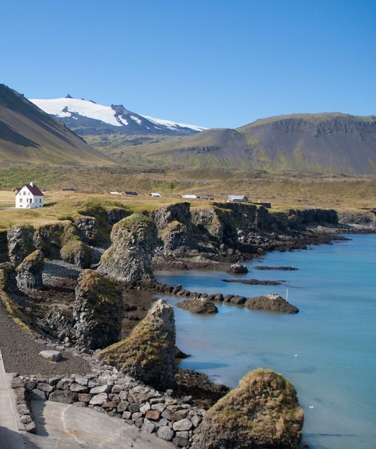A small house sits on a rocky shore, surrounded by natural landscape featuring mountains and a lake. The sky is clear, and the scene includes coastal landforms and a body of water.