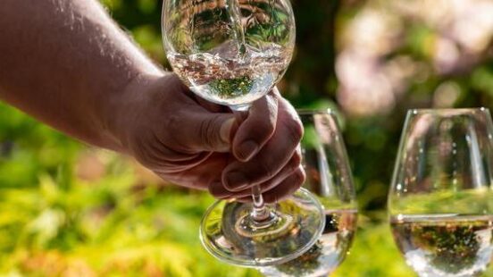 Pouring water into a wine glass with other glasses and greenery in the background.
