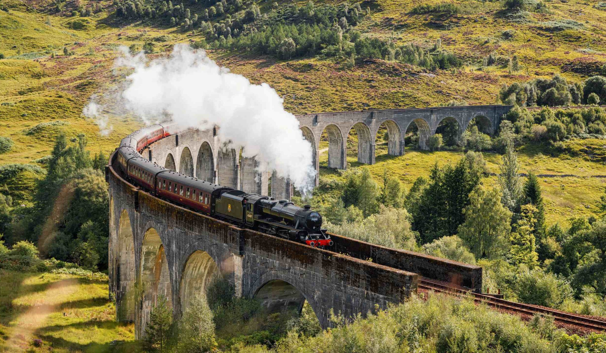 A steam train crosses a curved stone viaduct over a lush green valley in summer, a famous view along some luxury Europe rail tours.