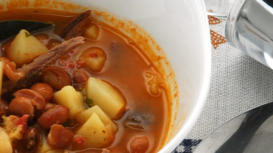 A bowl of hearty stew with beans, potatoes, and meat on a checkered tablecloth, with a spoon and empty glass.