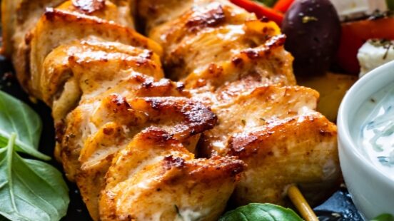Grilled chicken skewers with Greek salad and tzatziki sauce on a dark plate.