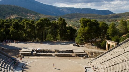Ancient amphitheater with stone seating surrounded by trees and mountains under a cloud-filled sky.