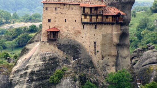 A historic monastery perched atop a tall, slender rock formation surrounded by greenery.