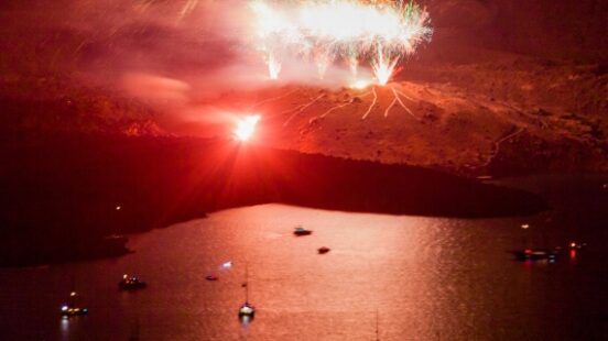 Fireworks over a reflective body of water with boats, and a bright flare near the shore.