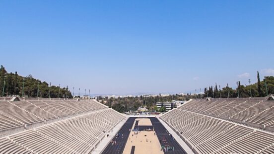 Empty ancient stadium with a running track, tiers of seats under a clear blue sky.