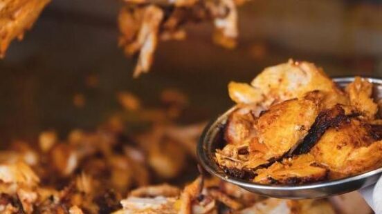 Close-up of a vendor carving pieces of roasted chicken from a vertical shawarma broiler.