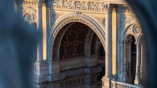 Sunlight illuminates the ornate archway of Galleria Vittorio Emanuele II in Milan, viewed through a blur.