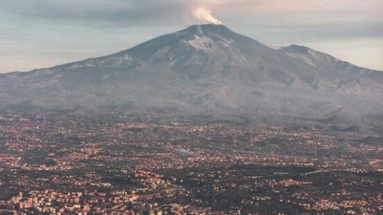 Aerial view of a city by the sea with a large, smoking volcano looming in the background.