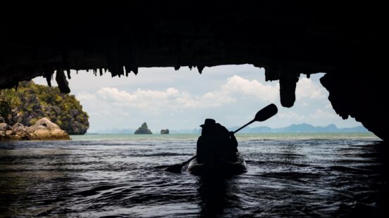 A kayaker paddling in the sea, silhouetted against a cave opening with distant islets.
