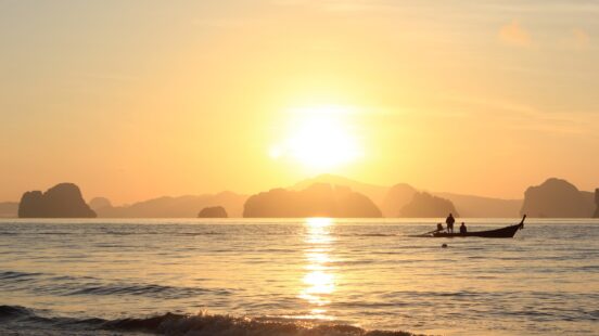 Sunset over the sea with silhouetted boat and people, against a backdrop of distant islands.
