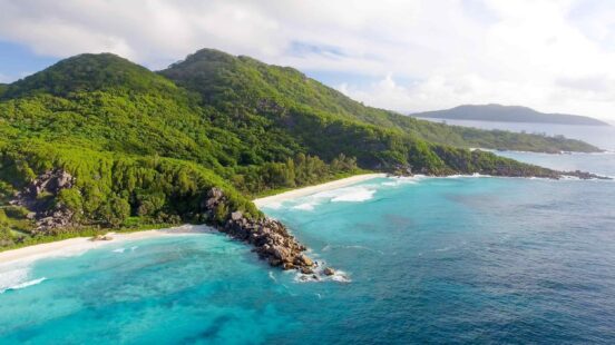 Aerial view of lush green mountains meeting a turquoise bay and sandy beach, with gentle waves lapping at the shore under a blue sky.