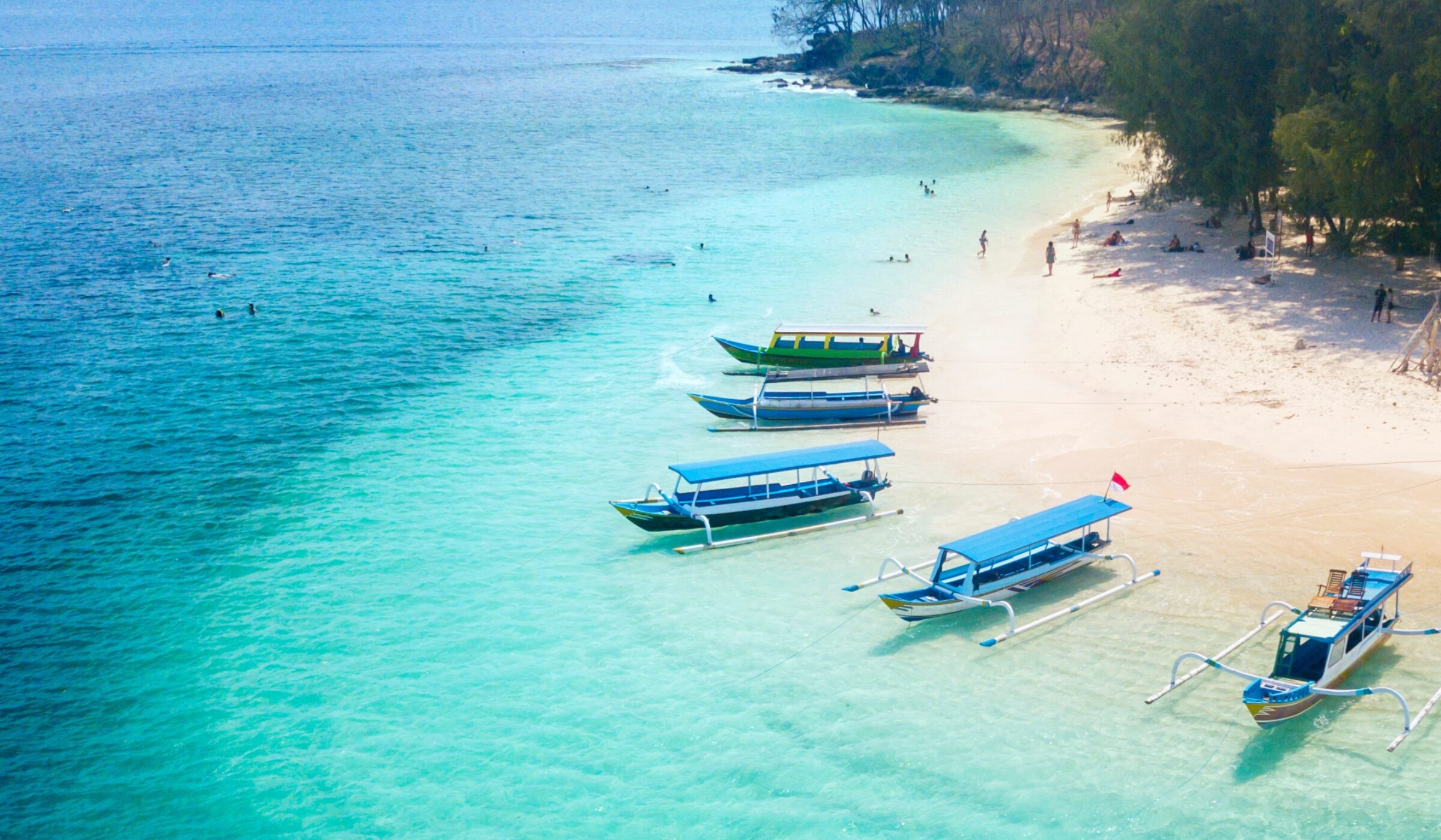 Colorful boats anchored on a tranquil beach with clear turquoise water and people enjoying the sun and water activities.