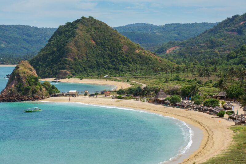 Scenic beach view with turquoise water, sandy shores, lush greenery, and hills in the background, dotted with small huts.