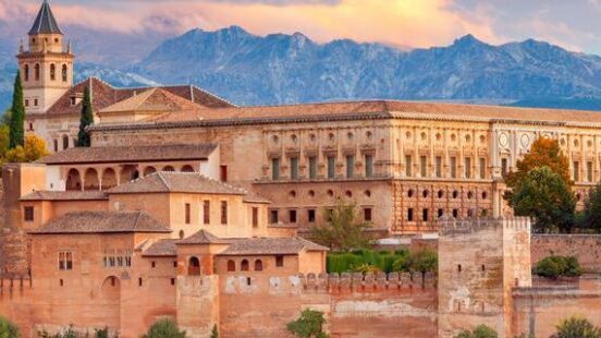 The vast, historic Alhambra palace and fortress complex in Granada, Spain, surrounded by green trees, with mountains and a colorful sunset sky.