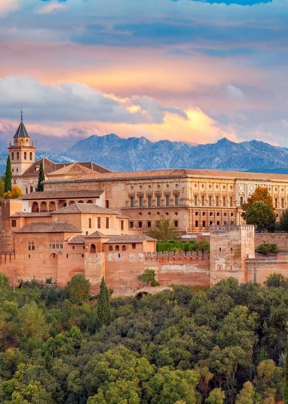 The vast, historic Alhambra palace and fortress complex in Granada, Spain, surrounded by green trees, with mountains and a colorful sunset sky.