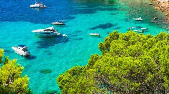 Several boats anchor in a turquoise-blue cove in the Balearic Islands with a rocky cliff, green trees, and buildings above.