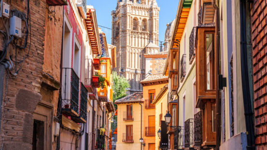 A narrow, uphill cobblestone street in Toledo, Spain, with old buildings on both sides, leading to a tall cathedral spire.