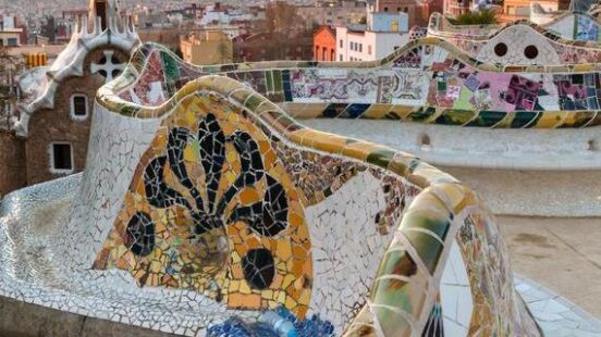 A colorful, winding mosaic-tiled bench at Park Güell in Barcelona, Spain, with the cityscape and ocean visible under a pastel sky.