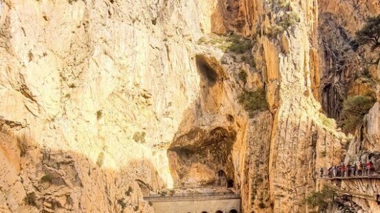 Steep, towering canyon walls feature a recessed arched structure and a narrow walkway clinging to the right side.