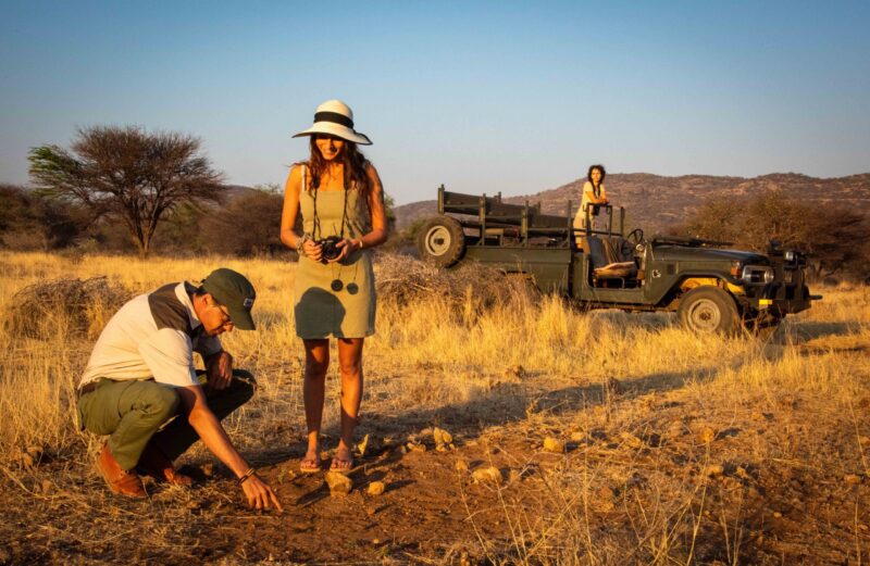 A safari guide squats to point at the ground while a woman with a camera watches near a parked off-road vehicle.