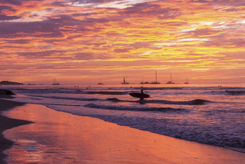 A surfer silouetted against a brilliantly colored sunset as he comes in with his surfboard on Playa Tamarindo, Guanacaste, Costa Rica