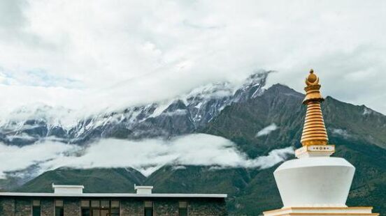 A white Buddhist stupa in front of a modern stone building and large, cloud-covered mountains.
