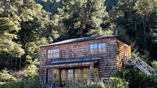 A rustic wooden cabin with large glass doors nestled at the base of a forested mountain.
