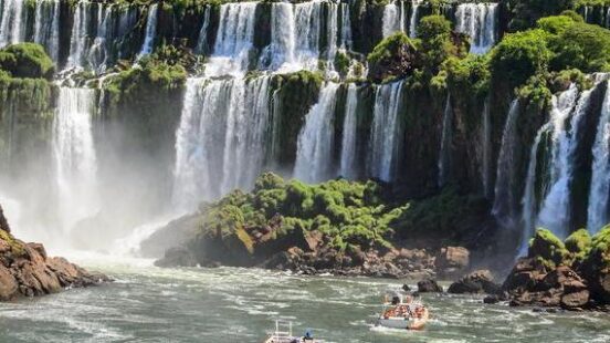 Two white tour boats approaching the base of massive, multi-tiered waterfalls under a bright blue sky.