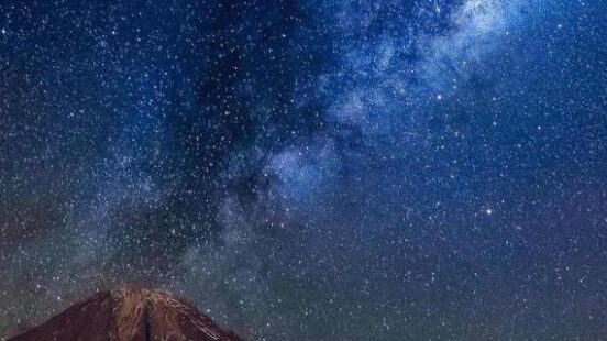 A brilliant view of the Milky Way stars in a blue and purple sky over a dark mountain peak.