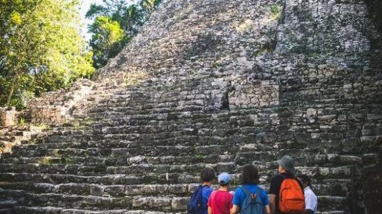 Tourists standing at the bottom of a tall, ancient stone step pyramid in a forested archaeological site.