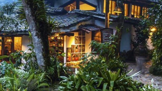 A stone water fountain in a lush garden leading to a glowing mountain villa at dusk.